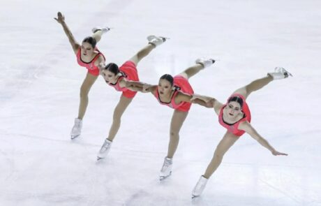 Quatre patineuses synchronisées en costume corail, bras liés, jambe tendue